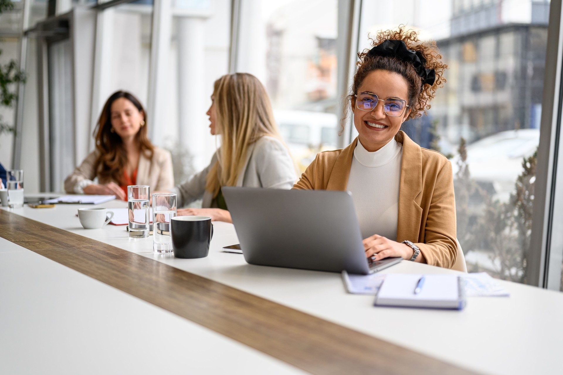 Confident businesswoman in eyeglasses working online over laptop while sitting with colleagues in board room